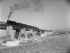 Composición de viajeros remolcada por locomotora a vapor a su paso por el puente metálico sobre e...