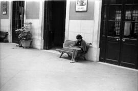 Mujer sentada en un banco de una estación sin identificar, posiblemente francesa, leyendo una rev...